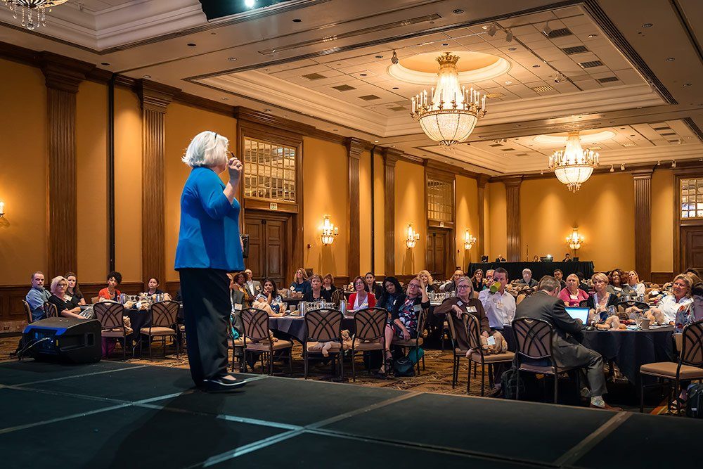Keynote speaker on stage in a hotel ballroom, with the audience at round tables filling the room behind